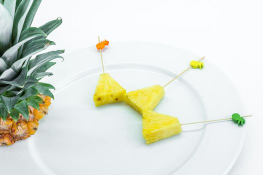 Pineapple skewers peeled and cut into triangles with chopsticks decorated on white plate with pineapple leaves on white background