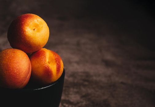 three red and yellow apricots with water drops in a black bowl with soft side lighting on dark spotty background