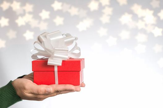 Hands of a woman in green sweater with red gift and white ribbon on neutral background with bokeh of stars