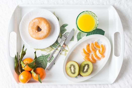 Image from above of a tray with a breakfast donut with mandarin kiwi and orange juice on a white bed with natural light