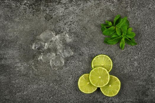 Close up heap of fresh green mint leaves, lime slices and ice cubes on grunge gray stone table surface to prepare mojito cocktail, elevated top view, directly above