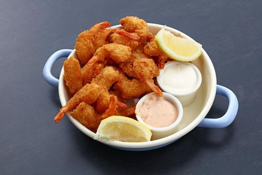 Close up portion of battered and breaded shrimps with dipping sauces served in cooking pot over blue table copy space, high angle view