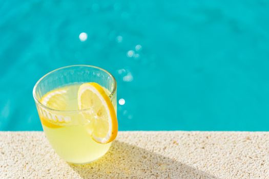 lemonade in transparent glass with lemon slice on the edge of the pool with turquoise blue water background in horizontal format