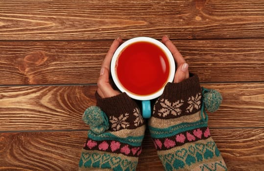 Close up two woman hands hold and hug big full cup of black tea or red fruit tea over brown wooden table, elevated top view, directly above