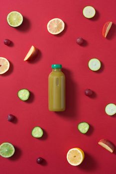 bottle of green fruit and vegetable juice in the center of the image surrounded by pieces of various colored fruits and vegetables on a red background