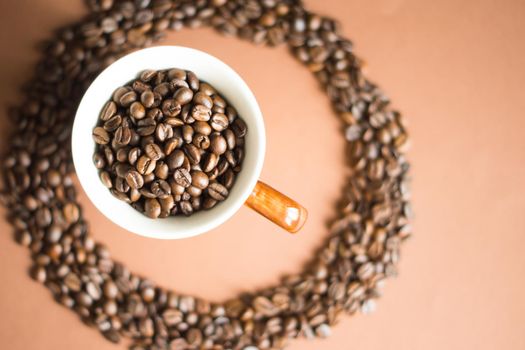 White cup with brown handle full of coffee beans picked from above with light brown background and circle of coffee beans