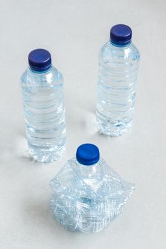 three plastic bottles with blue caps of mineral water one crushed empty to recycle first and two smaller ones standing behind on gray background