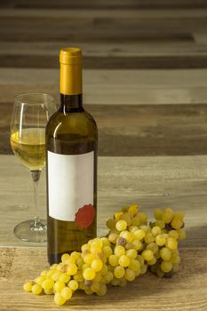 vertical composition of bottle of white wine with white label glass and white grapes with background of wooden boards of various shades of brown and soft light