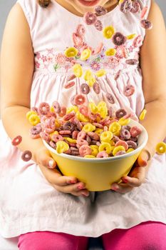 colored cereal rings falling into a yellow bowl held by a girl with both hands wearing a pink dress