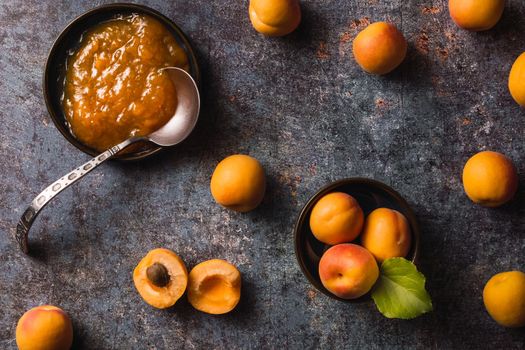 picture from above of apricots scattered around the scene three grouped in a bowl with a green leaf one cut in half and a bowl full of apricot jam with an old spoon