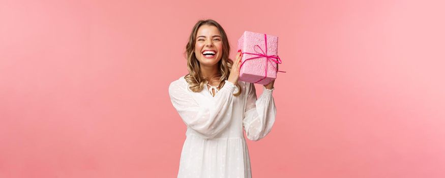 Holidays, celebration and women concept. Portrait of happy charismatic blond girl shaking gift box wondering whats inside as celebrating birthday, receive b-day presents, pink background.