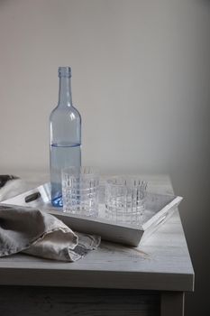 A blue transparent water bottle and two ribbed glasses on a white wooden tray on the kitchen table.