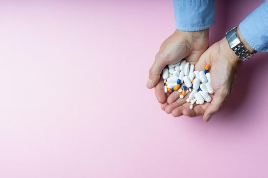 Caucasian man hands with blue shirt silver wristwatch holding handful of medications in tablets and capsules in varied shape and color size on pink background with side lighting