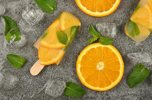 Close up two fruit ice cream popsicles with fresh orange slices, green mint leaves and ice cubes on gray table surface, elevated top view, directly above