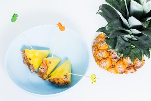 Pineapple skewers peeled and cut into triangles with chopsticks decorated inside a blue bowl with white background and pineapple leaves