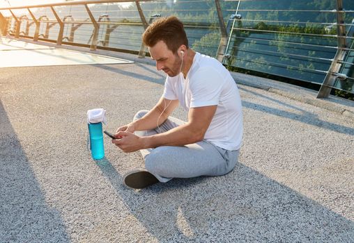 Middle aged handsome Caucasian sportsman wearing earphones sitting on the city bridge and checking his phone while relaxing after morning cardio workout and using a fitness tracking mobile application