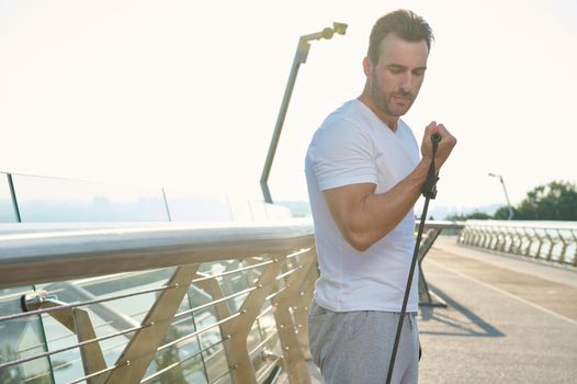 Handsome Caucasian middle aged man, athlete during outdoor workout with rubber resistance band. A middle-aged sportsman is training with an elastic expander on a glass city bridge at dawn