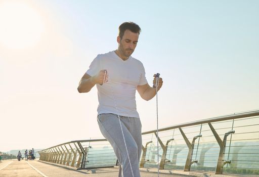 Handsome middle aged sportsman, muscular build Caucasian middle aged man training with jumping rope on a city bridge treadmill early in the morning on a beautiful sunny summer day. Keep your body fit