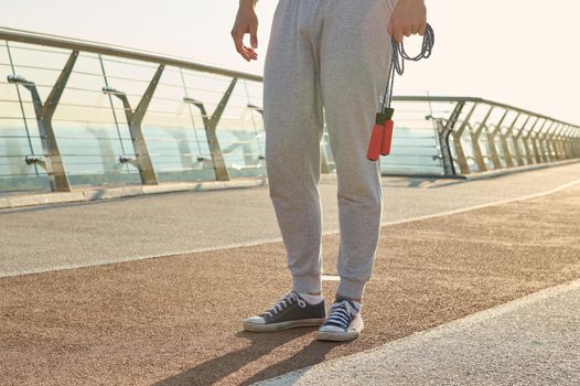 Cropped view of a sportsman holding a jumping rope while standing on a treadmill during a morning cardio workout. Close-up