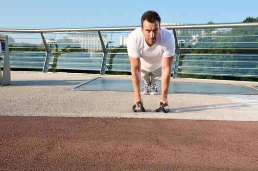 Muscular handsome attractive middle aged Caucasian sportsman, determined athlete doing push ups from the floor during an intense outdoor workout on an urban glass bridge on a warm early summer day