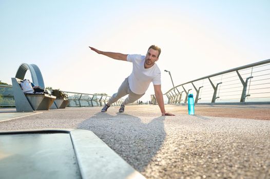 Active muscular European middle aged man, athlete, sportsman doing push-ups from the floor, alternately raising one arm during an outdoor workout at dawn on the city bridge