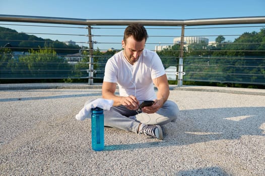 Middle aged European man, athlete using fitness tracking app on smartphone. Determined sportsman checking his phone , sitting on the city bridge and relaxing after outdoor heavy workout