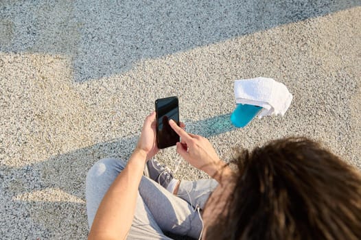 Top view of an European sportsman wearing earphones, holding a mobile phone and pointing his finger on a black blank screen with copy space. Athlete swiping on mobile application checking heart beat