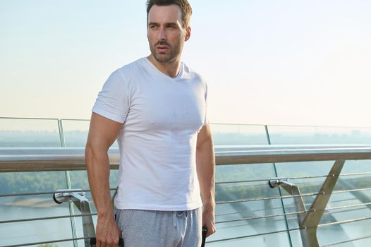 Determined middle aged muscular build Caucasian man, athlete, sportsman exercising with elastic rubber bands outdoor, looking aside standing on the city bridge at dawn