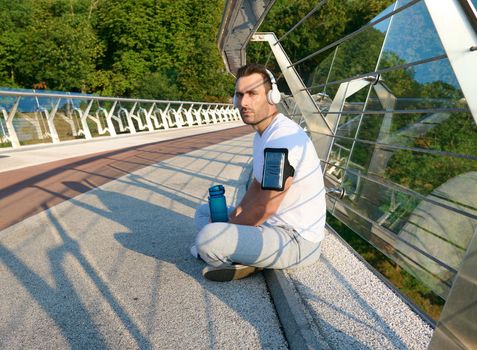 Confident relaxed muscular middle aged European man, athlete in sportswear and headphones, listening to the music while relaxing on the urban glass bridge after morning workout on a summer sunny day