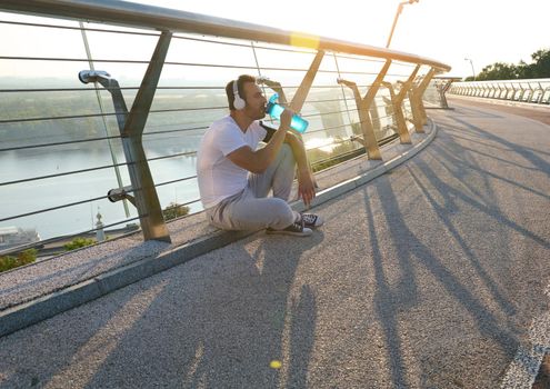 Handsome sportsman with headphones drinking water and relaxing after morning outdoor workout, sitting on the glass city bridge with sunbeams falling on the bridge treadmill. Enjoy sport in summer days