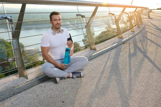 Handsome European muscular man, determined athlete, sportsman in headphones relaxing after workout outdoors at dawn, holding water bottle and sitting on glass city bridge in summer
