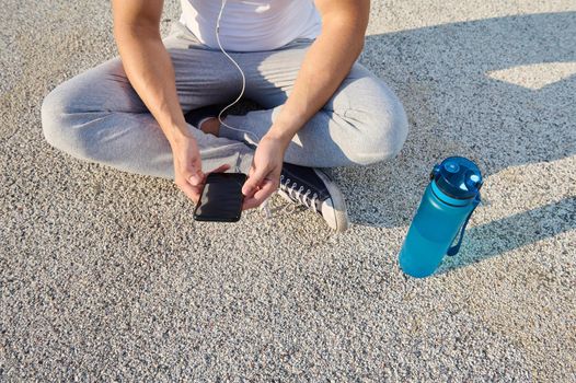Overhead view of an athlete wearing earphones sitting on the city bridge and checking his phone while relaxing after morning cardio workout and using a fitness tracking mobile application
