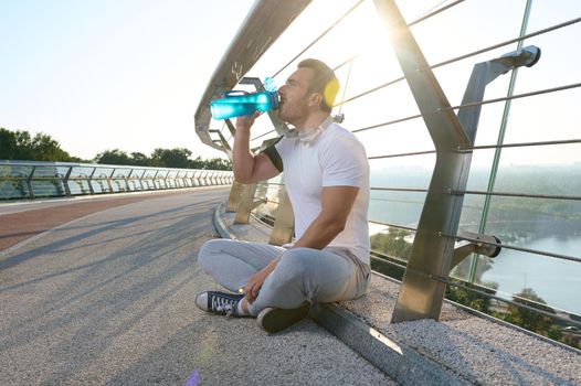 Side portrait of handsome attractive European man, athlete, muscular sportsman drinking water and relaxing after a heavy workout on a glass city bridge. The sunbeams of the early morning fall on him.