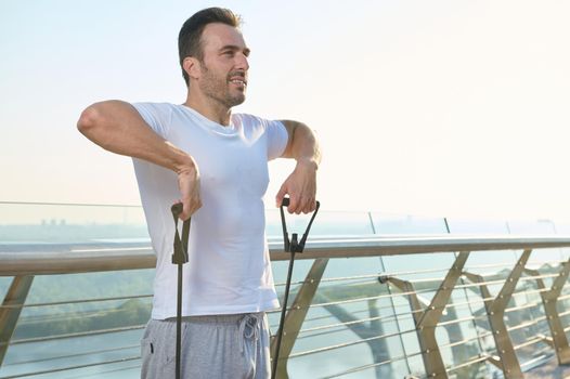 European young sportsman, athlete during outdoor workout with rubber resistance band. Middle-aged athlete doing exercises on shoulders and arms using an elastic expander on glass city bridge at dawn