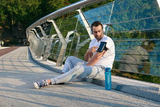 Attractive muscular man, athlete in headphones sits on a city bridge and checks a mobile application with monitoring heart rate and calories burned during a workout on his phone in a smartphone holder