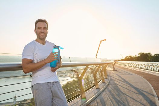 Confident athlete, Caucasian man with muscular aesthetic body in sportswear holds a bottle of water, refreshes himself after workout and smiles looking at camera, standing on an urban bridge at dawn