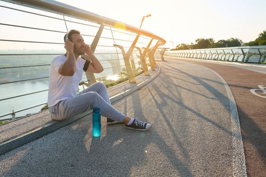 Attractive relaxed young muscular man athlete putting on headphones and listening to music, sitting on a bridge and enjoying the rest after morning workout. Beautiful sun rays falling on the treadmill