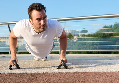 Close-up of a determined handsome middle aged Caucasian man, athlete exercising outdoor, pumping muscles while performing push-ups training on the urban bridge on a beautiful summer day