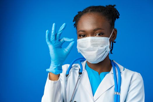 Portrait happy African American female doctor showing ok sign in blue studio, close up
