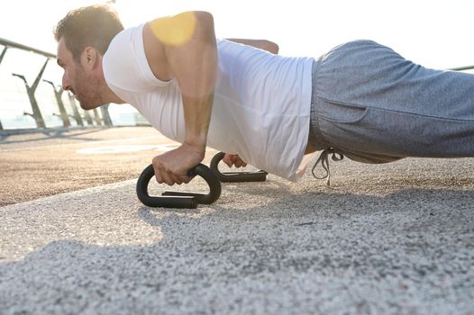 Close-up portrait of a handsome middle aged European man, muscular build athlete, sportsman doing push ups exercises during an outdoor workout on the city bridge early in the morning at dawn