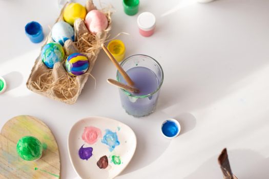 On a white table, in daylight, there are brushes in a glass of water, multi-colored eggs in a tray, jars of paint. Top view. Copy space