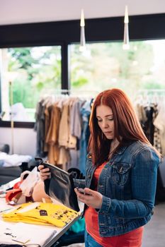 young woman with red hair, enjoying a shopping day, leisure concept. looking at price tags of clothes to buy, in a fashion shop. shopping concept. Natural light, sun rays, display with clothes, clothes rack, customers in background walking, clothes, Vertical view. customer with blue denim jacket, red t-shirt and jeans.