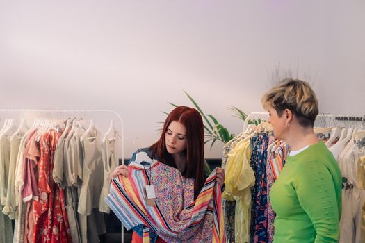 young woman customer advised to buy a dress, by a saleswoman expert in clothes, in a fashion shop. shopping concept. leisure concept. business owner selling. Natural light, sun rays, display with clothes, horizontal view, copy space. customer in blue denim jacket, saleswoman in green t-shirt.