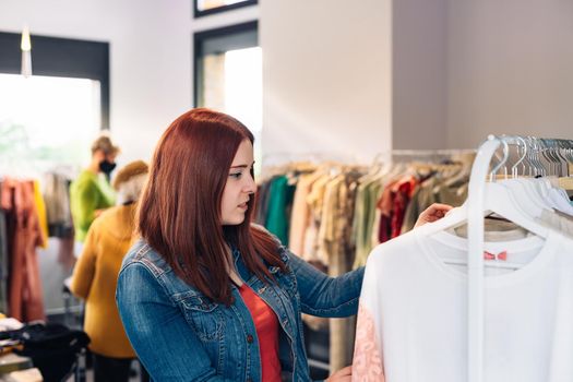 Young woman with red hair, happy and enjoying a day of shopping, choosing a white t-shirt from a hanger, shopping in a fashion shop shopping concept.natural light, sun rays, display with clothes, clothes rack, shop window, clothes, Horizontal.