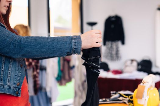 hands of a young woman, enjoying a shopping day, choosing a t-shirt to buy, in a fashion shop. shopping concept. Natural light, sun rays, display with clothes, clothes rack, customers in background walking, clothes, Horizontal. customer with blue denim jacket, red t-shirt and jeans.