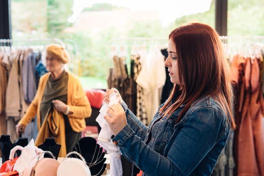 young red-haired woman looking at clothes in a fashion shop, deciding what to buy, shopping concept. leisure concept. background customers walking. Natural light, sunbeams, display, clothes rack, clothes, horizontal view, copy space. customer in blue denim jacket, red t-shirt.