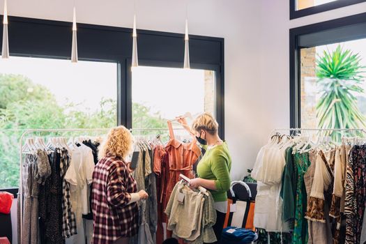 mature fashion saleswoman advising her customers, in small fashion shop. fashion shop customers shopping. shopping concept. natural light, sun rays, display with clothes, clothes rack, shop window, clothes, horizontal, saleswoman dressed in green t-shirt, customer dressed in striped shirt.