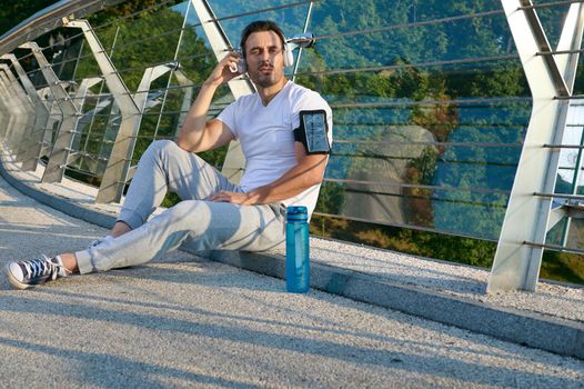 Confident relaxed handsome Caucasian man, athlete in sportswear, putting on headphones and listening to the music while relaxing on the urban glass bridge after morning workout on a summer sunny day