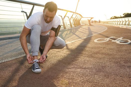 Middle aged Caucasian man athlete jogger tying shoelaces on treadmill during morning jog at dawn. Cardio workout, jogging, running, outdoor sports training, Motion, endurance healthy lifestyle concept