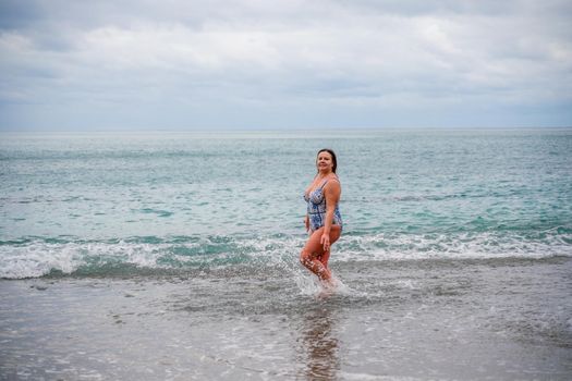 A plump woman in a bathing suit enters the water during the surf. Alone on the beach, Gray sky in the clouds, swimming in winter